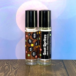 May include: Two 10 ml roll-on bottles of essential oil blend. The bottle on the left has a brown label with a cartoon image of soot sprites from the movie 'My Neighbor Totoro'. The bottle on the right has a black label with the text 'Soot Sprites' and 'Embers + Smoke + Leaves Fragrance'.
