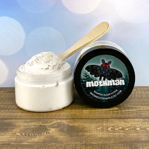 May include: A white whipped sugar scrub in a clear jar with a black lid. The jar is labeled "mothman whipped sugar scrub snowscutsoops.etsy.com". The label features a black moth with red eyes against a green background.