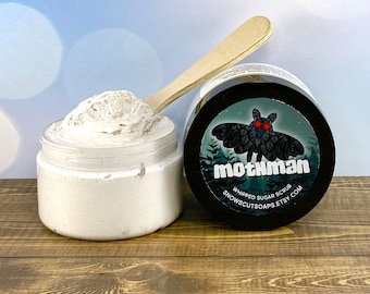May include: A white whipped sugar scrub in a clear jar with a black lid. The jar is labeled "mothman whipped sugar scrub snowscutsoops.etsy.com". The label features a black moth with red eyes against a green background.