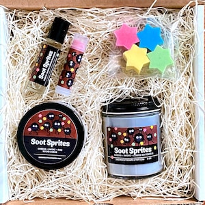May include: A gift box filled with a gray soy candle, sugar scrub, lip balm, and essential oil, all featuring a black and white illustration of soot sprites. The box is filled with shredded paper and includes a small bag of four star-shaped soaps in pink, blue, yellow, and green.