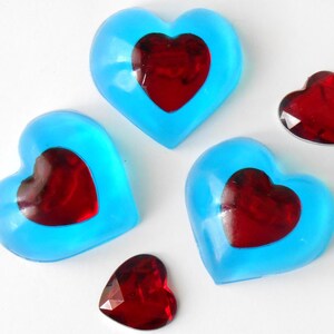 Peut inclure: Quatre savons en forme de cœur, trois sont bleu clair avec un cœur rouge à l'intérieur, et un est un cœur rouge. Les savons sont sur une surface blanche.