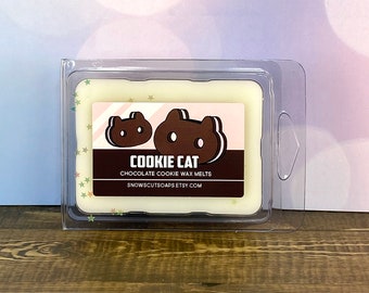 May include: A white wax melt in a clear plastic container with a label that says "COOKIE CAT CHOCOLATE COOKIE WAX MELTS SNOWSCUTSOAPS.ETSY.COM". The label has a cartoon image of a brown cat with a cookie on its head.