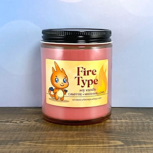 May include: A red soy candle with a black lid and a label that says "Fire Type" with a cartoon fire-type Pokemon on it. The label also says "soy candle" and "Campfire + Marshmallows".