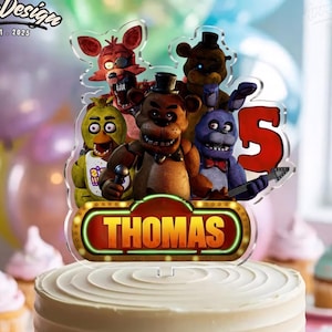 May include: A cake topper featuring characters from a popular video game franchise, with the name "THOMAS" and the number "5". The topper is placed on a frosted cake, surrounded by cupcakes and balloons.