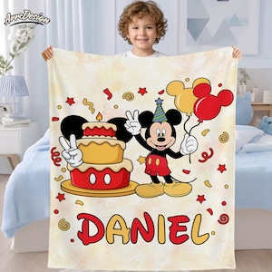 May include: A personalized blanket featuring Mickey Mouse holding balloons and a birthday cake. The blanket has a light yellow background with red and yellow accents, confetti, and the name "DANIEL" in red and yellow lettering. The blanket is held by a child.