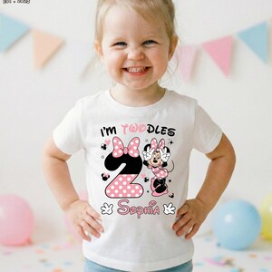 May include: White toddler t-shirt with a Minnie Mouse design. The design includes the text "I'm Twoodles" and "Sophia", along with a large number "2" in pink polka dots. The shirt is worn by a smiling child.