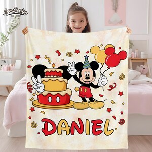 May include: A cream-colored blanket featuring a cartoon Mickey Mouse holding balloons and a birthday cake. The blanket has the name "DANIEL" in red and yellow, with confetti and star accents. The blanket is held up by a young girl.
