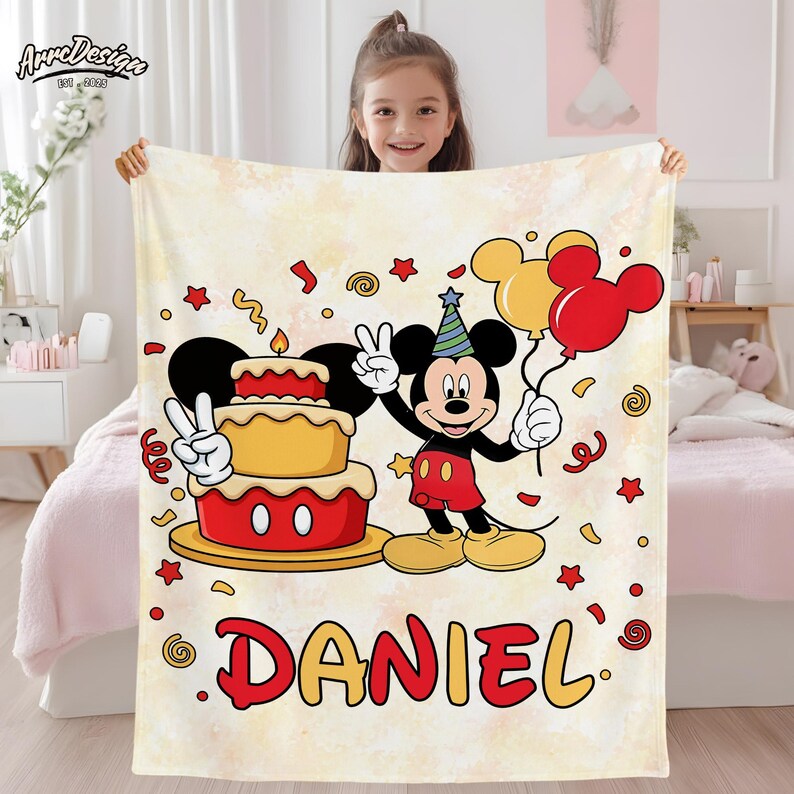 May include: A cream-colored blanket featuring a cartoon Mickey Mouse holding balloons and a birthday cake. The blanket has the name "DANIEL" in red and yellow, with confetti and star accents. The blanket is held up by a young girl.