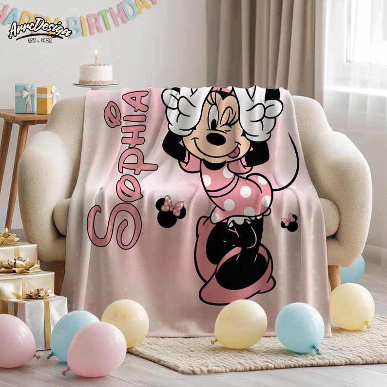 May include: A pink blanket with a Minnie Mouse design and the name "Sophia". The blanket has a polka dot pattern and is decorated with balloons and a birthday banner. The blanket is on a sofa.