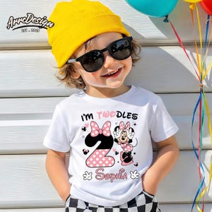 May include: A white t-shirt with the text "I'm Twoodles" and the number "2" in pink polka dots. The shirt features a Minnie Mouse graphic and the name "Sophia". The child is wearing a yellow beanie and sunglasses.