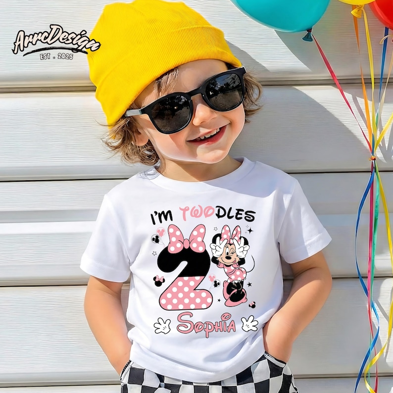 May include: A white t-shirt with the text "I'm Twoodles" and the number "2" in pink polka dots. The shirt features a Minnie Mouse graphic and the name "Sophia". The child is wearing a yellow beanie and sunglasses.