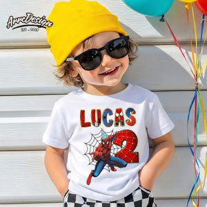 May include: White t-shirt featuring the name "LUCAS" in colorful letters and a graphic of Spider-Man with a "2" for a birthday. The child is wearing a yellow beanie and sunglasses. The image includes balloons.