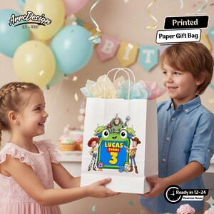 May include: A white paper gift bag featuring a colorful graphic of Toy Story characters surrounding a green frog-shaped frame with the text Lucas Turns 3. The bag has white paper handles and contains light-colored tissue paper at the top opening.