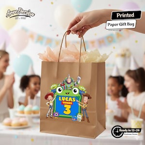 May include: A brown paper gift bag featuring a colorful graphic of Toy Story characters including Buzz Lightyear, Woody, Jessie, and green aliens surrounding a green frog shape with the text Lucas Turns 3. The bag has brown paper handles.