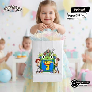 May include: A white paper gift bag featuring a colorful graphic of Toy Story characters surrounding a green frog-shaped frame with the text Lucas Turns 3. The bag is filled with pastel-colored tissue paper and has white twisted paper handles.