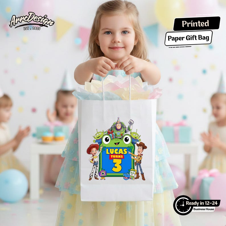 May include: A white paper gift bag featuring a colorful graphic of Toy Story characters surrounding a green frog-shaped frame with the text Lucas Turns 3. The bag is filled with pastel-colored tissue paper and has white twisted paper handles.