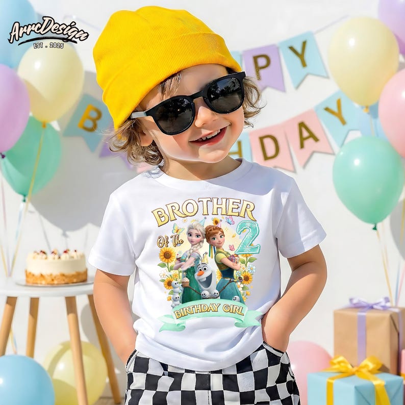 Pu&ograve; includere: Una maglietta bianca con la scritta "Brother of the Birthday Girl" e una grafica di due personaggi animati, girasoli e il numero 2. Il bambino indossa un berretto giallo e occhiali da sole.