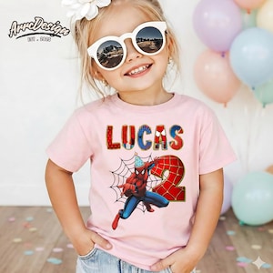 May include: A light pink t-shirt featuring the name "LUCAS" in a colorful, textured font, and a graphic of Spider-Man with a party hat and the number "2". The shirt also has a spiderweb design. The ArreDesign logo is in the top left corner.