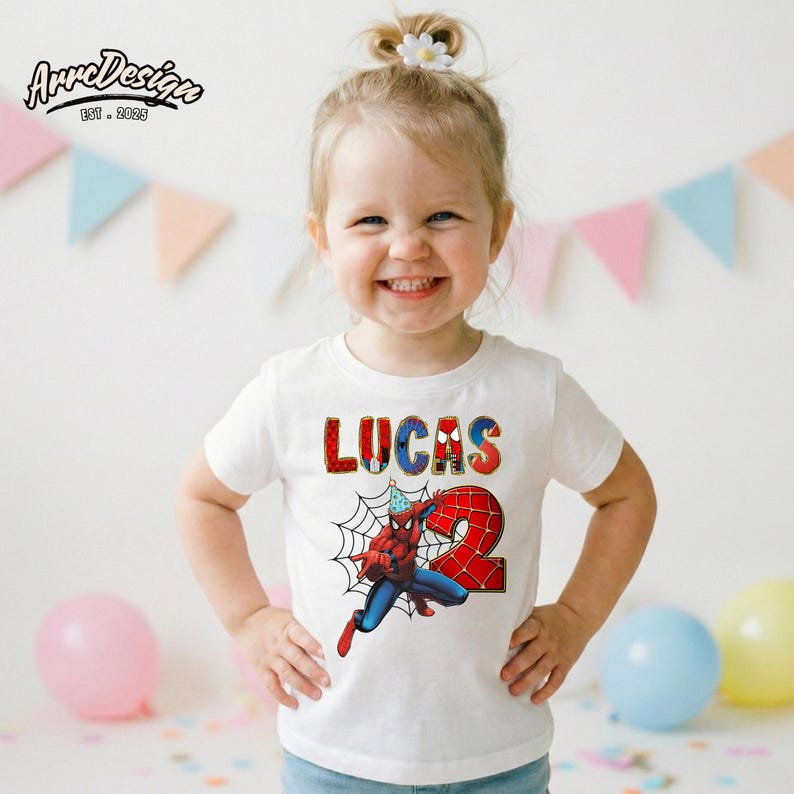 May include: White t-shirt featuring a colorful graphic of Spider-Man with the name "LUCAS" and the number "2". The design includes a spider web and a birthday hat. The shirt is worn by a young child.