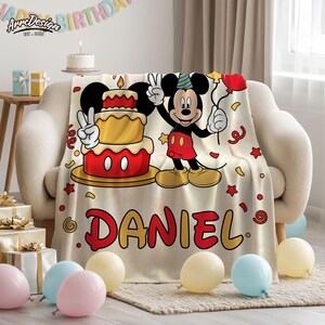 May include: A beige blanket featuring a cartoon Mickey Mouse design with a birthday cake and balloons. The blanket has the name "DANIEL" in red letters. The blanket is draped over a beige sofa, with balloons and gifts in the foreground.