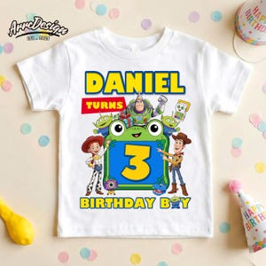 May include: A white short-sleeve t-shirt featuring a colorful graphic with the text DANIEL TURNS 3 BIRTHDAY BOY. The design includes cartoon characters Buzz Lightyear, Woody, Jessie, and green aliens surrounding a large yellow number 3.