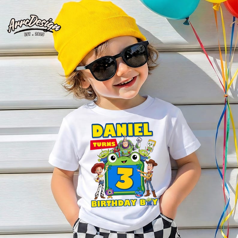 May include: A white short-sleeved t-shirt featuring a colorful graphic print with the text DANIEL TURNS 3 BIRTHDAY BOY. The design includes cartoon characters from Toy Story surrounding a green frog-shaped frame with the number 3 inside.