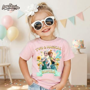 Pu&ograve; includere: Una bambina indossa una maglietta rosa con la scritta "TWO & MAGICAL 2" e il nome "SOPHIA". La maglietta presenta una grafica di personaggi animati e girasoli. Indossa occhiali da sole bianchi e un fiore bianco tra i capelli.