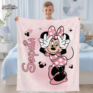 May include: Pink fleece blanket featuring Minnie Mouse with a polka dot bow and dress. The blanket has the name "Sophia" in pink script, along with Minnie Mouse illustrations and polka dots. The blanket is held by a child.