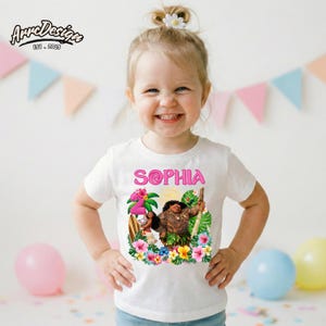 May include: White t-shirt featuring a colorful graphic of a cartoon character with the name "SOPHIA" in pink. The design includes tropical flowers and foliage. The child is smiling and wearing the shirt.