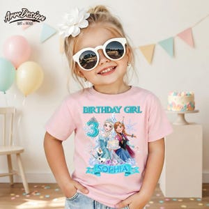 May include: A light pink short-sleeved t-shirt featuring a graphic print of two animated characters, a large number 3, and the text "BIRTHDAY GIRL" and "SOPHIA". The design includes snowflake motifs and a cake illustration in shades of blue and purple.