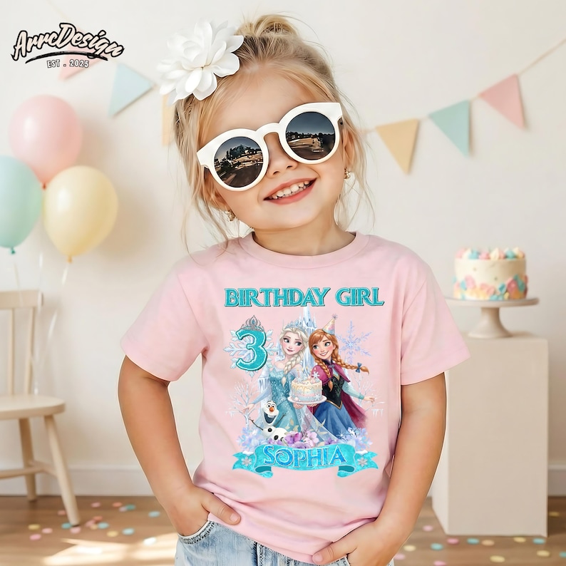 May include: A light pink short-sleeved t-shirt featuring a graphic print of two animated characters, a large number 3, and the text "BIRTHDAY GIRL" and "SOPHIA". The design includes snowflake motifs and a cake illustration in shades of blue and purple.