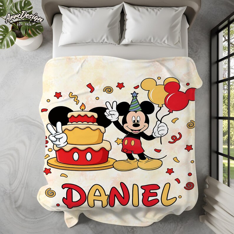 May include: A cream-colored blanket featuring a birthday theme with Mickey Mouse. The design includes a two-tiered cake, balloons, confetti, and the name "DANIEL" in red and yellow. Mickey is wearing a party hat.