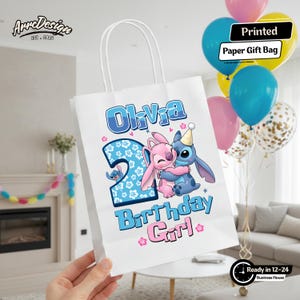 May include: White paper gift bag with the text "Olivia 2nd Birthday Girl" and a cartoon illustration of two characters hugging. The bag has white handles and is decorated with blue and pink accents. Balloons in various colors are in the background.