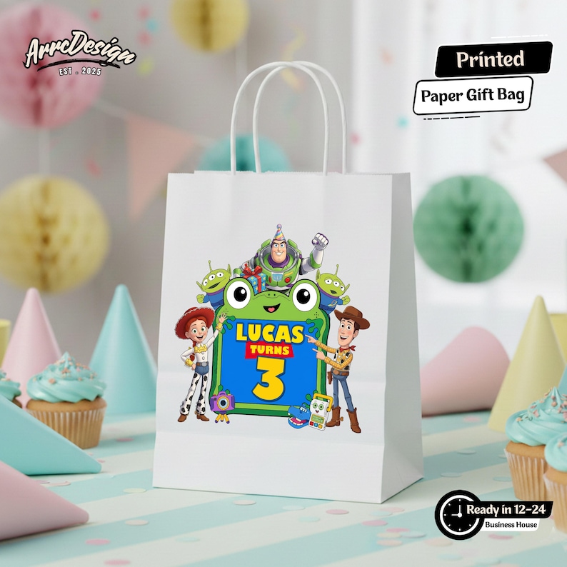 May include: A white paper gift bag featuring a colorful graphic of Toy Story characters including Woody, Jessie, Buzz Lightyear, and aliens surrounding a green frog shape with the text Lucas Turns 3. The bag has two white paper handles.