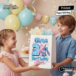 May include: A white paper gift bag with the text "Olivia" and "2 Birthday Girl" in blue and pink. The bag features a cartoon illustration of two characters hugging. The bag is held by two children.
