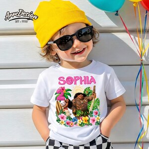 May include: White t-shirt with the name "Sophia" in pink, featuring a colorful graphic of characters and tropical flowers. The child is wearing a yellow beanie and sunglasses. The image also includes balloons.