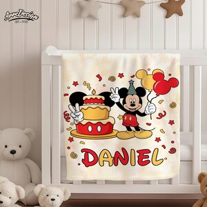 May include: A cream-colored baby blanket featuring a cartoon of Mickey Mouse holding balloons and a birthday cake. The name "DANIEL" is printed in red and yellow. The blanket is decorated with confetti and stars.