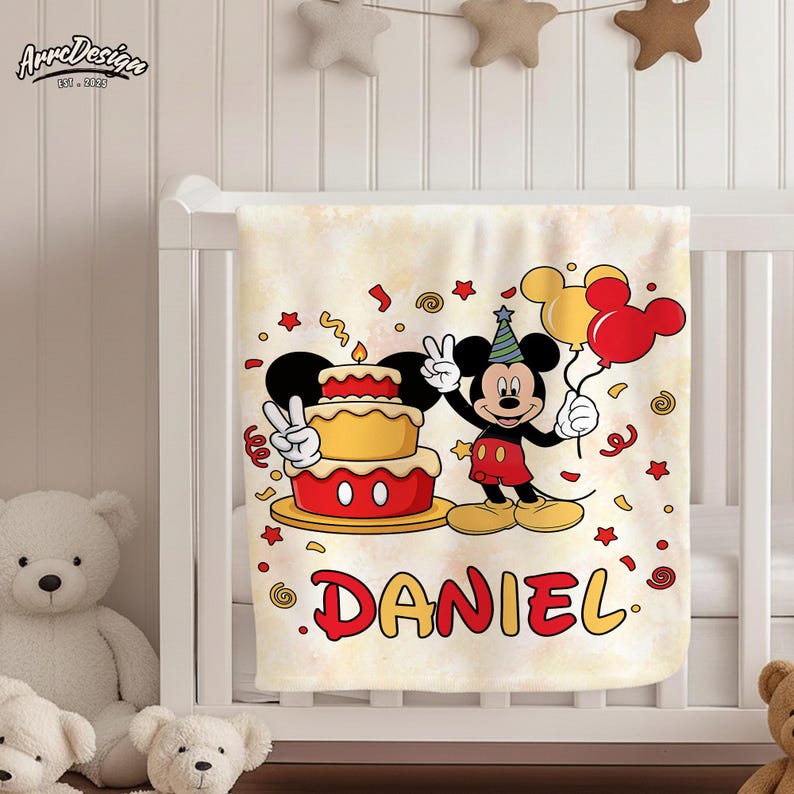 May include: A cream-colored baby blanket featuring a cartoon of Mickey Mouse holding balloons and a birthday cake. The name "DANIEL" is printed in red and yellow. The blanket is decorated with confetti and stars.