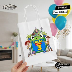 May include: A white paper gift bag featuring a colorful graphic of Toy Story characters including Buzz Lightyear, Woody, Jessie, and aliens surrounding a green frog-shaped frame with the text Lucas Turns 3 in blue and yellow lettering.