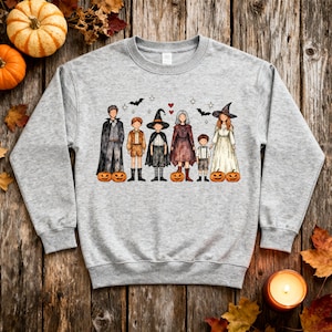May include: A heather gray crewneck sweatshirt featuring a watercolor-style illustration of six figures in various costumes, including witches and caped characters, surrounded by small bats, stars, hearts, and several orange jack-o'-lanterns.