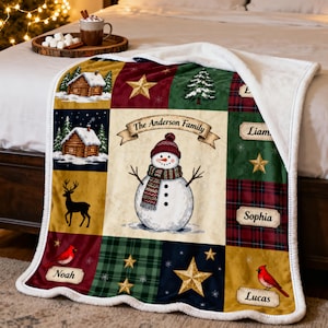 May include: A plush throw blanket featuring a central snowman wearing a red hat and scarf with the text The Anderson Family. The design includes panels with log cabins, deer, cardinals, stars, and name tags for Noah, Liam, Sophia, and Lucas.