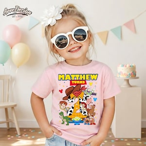 Puede incluir: Una ni&ntilde;a peque&ntilde;a con una camiseta rosa con un gr&aacute;fico colorido con personajes de la pel&iacute;cula Toy Story. La camiseta dice "MATTHEW TURNS" en texto amarillo y rojo. Tambi&eacute;n lleva gafas de sol blancas y una flor blanca en el pelo.