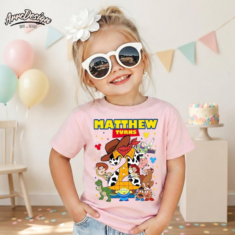 Puede incluir: Una ni&ntilde;a peque&ntilde;a con una camiseta rosa con un gr&aacute;fico colorido con personajes de la pel&iacute;cula Toy Story. La camiseta dice "MATTHEW TURNS" en texto amarillo y rojo. Tambi&eacute;n lleva gafas de sol blancas y una flor blanca en el pelo.