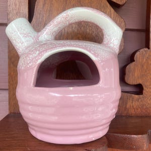 May include: A pink ceramic pitcher with a white glaze and a speckled pattern. The pitcher has a handle and a spout. It is a vintage style pitcher.