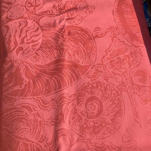 May include: Coral-colored fabric with a repeating pattern of stylized seashells and wave-like designs. The print features various shell shapes, including spirals and conch shells, in a lighter shade of coral. The fabric appears to be a textile, possibly for home decor or apparel.