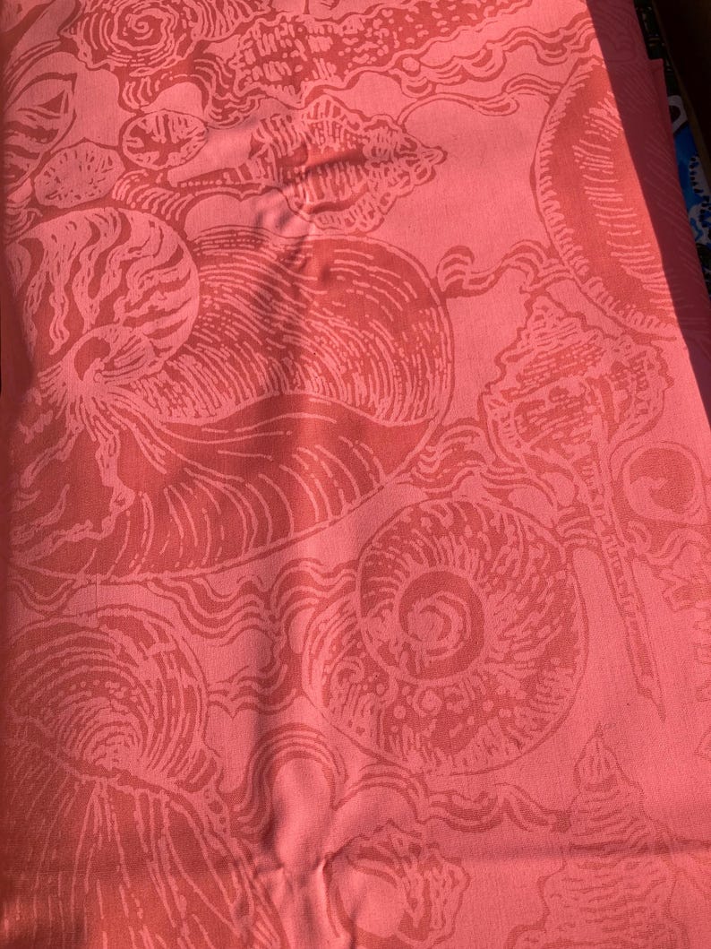 May include: Coral-colored fabric with a repeating pattern of stylized seashells and wave-like designs. The print features various shell shapes, including spirals and conch shells, in a lighter shade of coral. The fabric appears to be a textile, possibly for home decor or apparel.