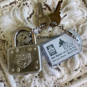 May include: A silver-toned metal Master padlock with a lion emblem and the number 77, attached to a ball chain with two keys and a rectangular plastic hotel key tag labeled Best Western Inn At The Park, Saint Louis, Missouri, 800-373-7501.