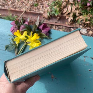 May include: A teal hardcover book with a white spine and a light brown interior. The book is lying on a blue wooden surface.