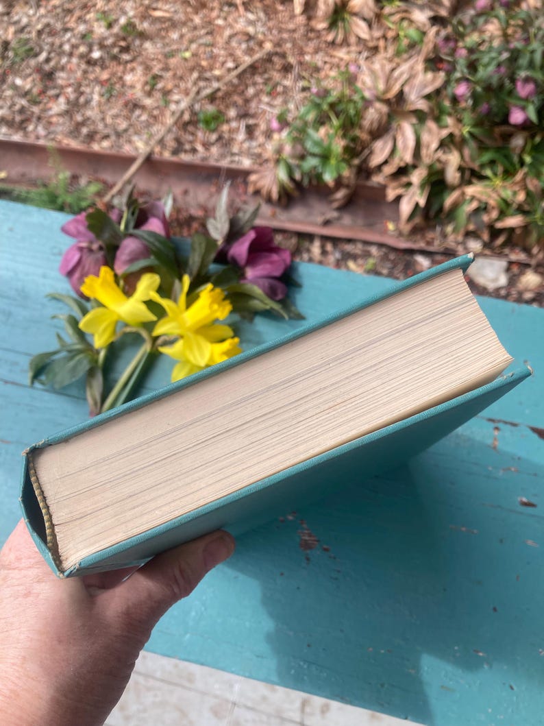 May include: A teal hardcover book with a white spine and a light brown interior. The book is lying on a blue wooden surface.