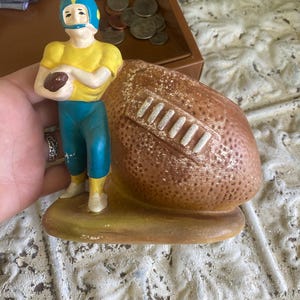 Puede incluir: Una figura de cerámica de un jugador de fútbol americano con camiseta amarilla, pantalones azules y casco azul con una franja amarilla. El jugador sostiene un balón marrón, junto a una maceta grande y texturizada con forma de balón.